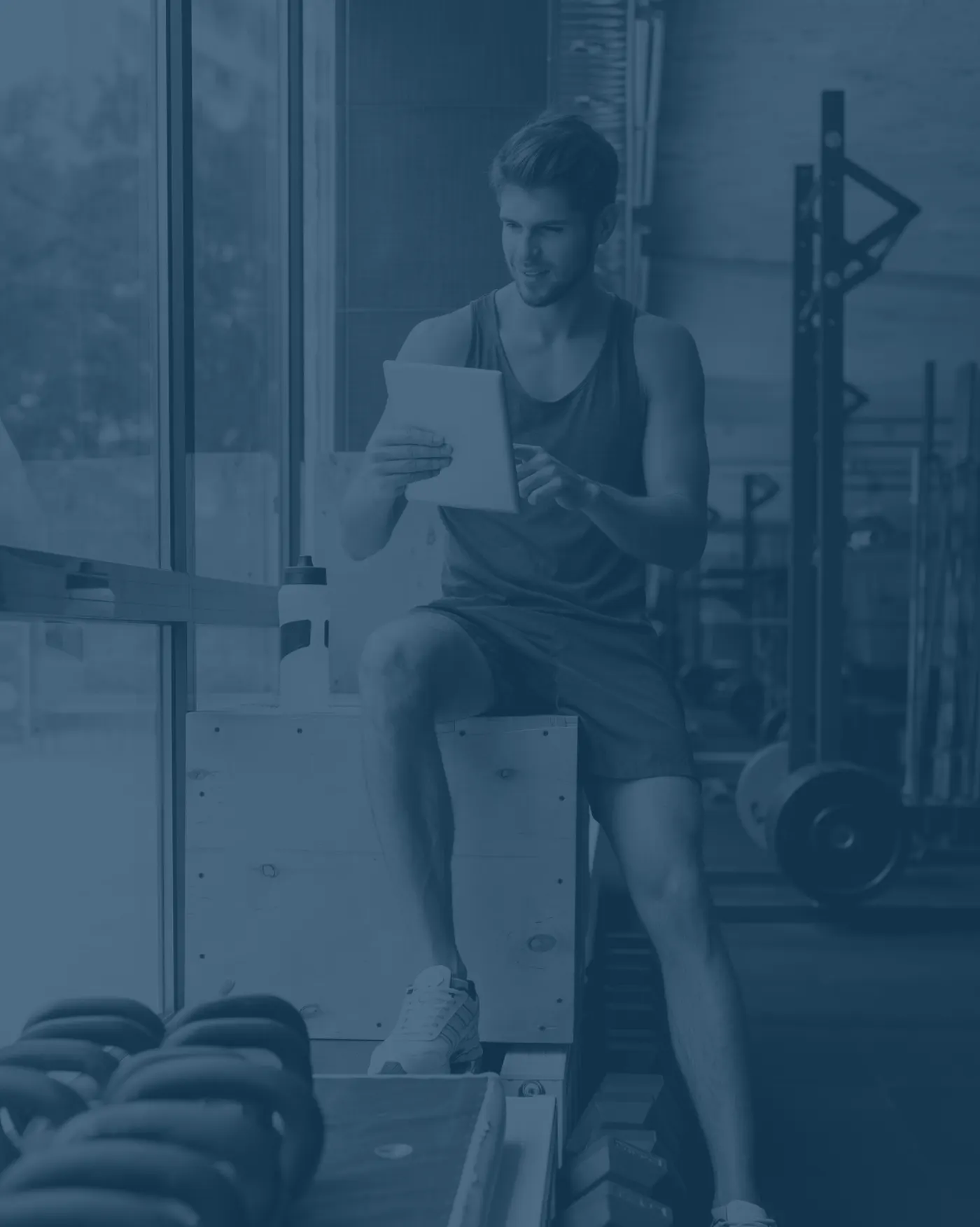 An athlete sits on a box in a gym, looking at a tablet.